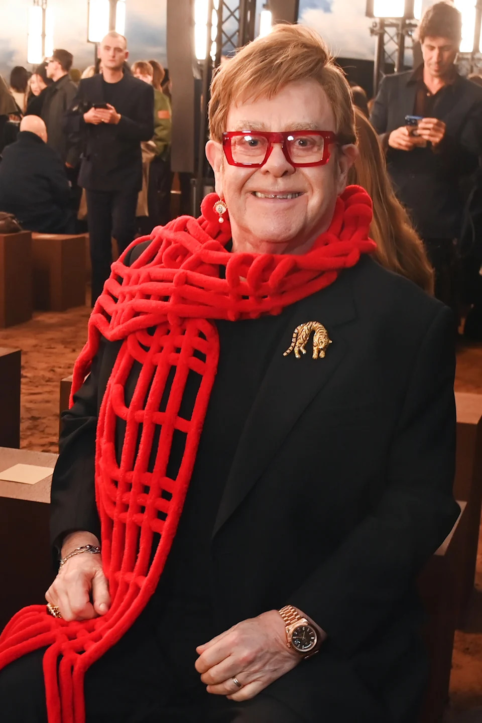 Person wearing a red scarf and glasses, seated at an event. They're dressed elegantly in a black outfit with jewelry and accessories