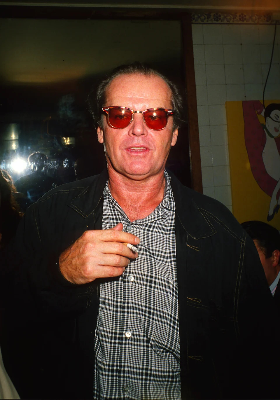 A person wearing sunglasses and a checkered shirt holds a cigarette. They wear a dark jacket and stand indoors