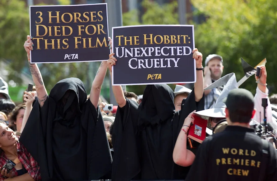 Protesters in hooded cloaks hold signs criticizing animal treatment during "The Hobbit" filming, surrounded by a crowd at an event