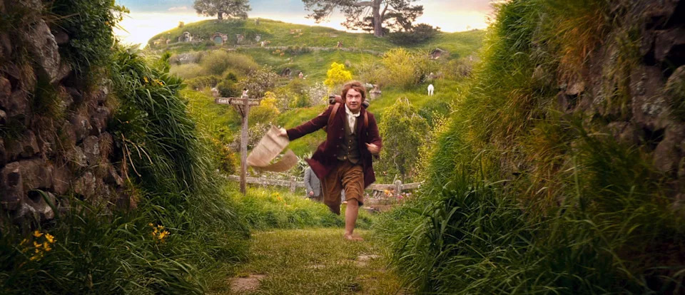 A person joyfully runs barefoot through a grassy path in a lush, picturesque landscape with hills and small hobbit-like homes in the background
