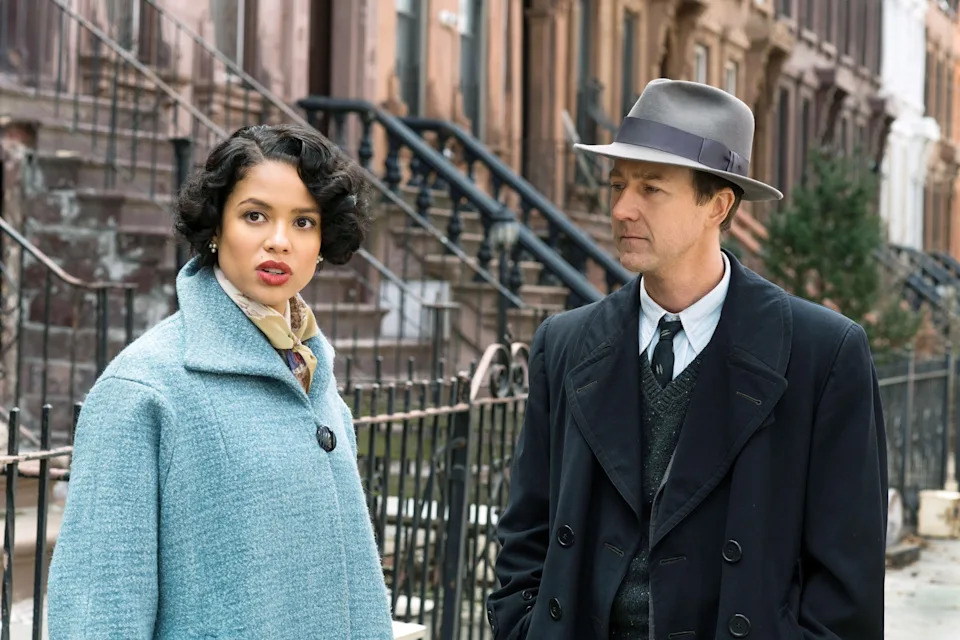Two people in vintage-style clothing stand outside by a wrought-iron fence. The woman wears a classic coat; the man wears a fedora and an overcoat