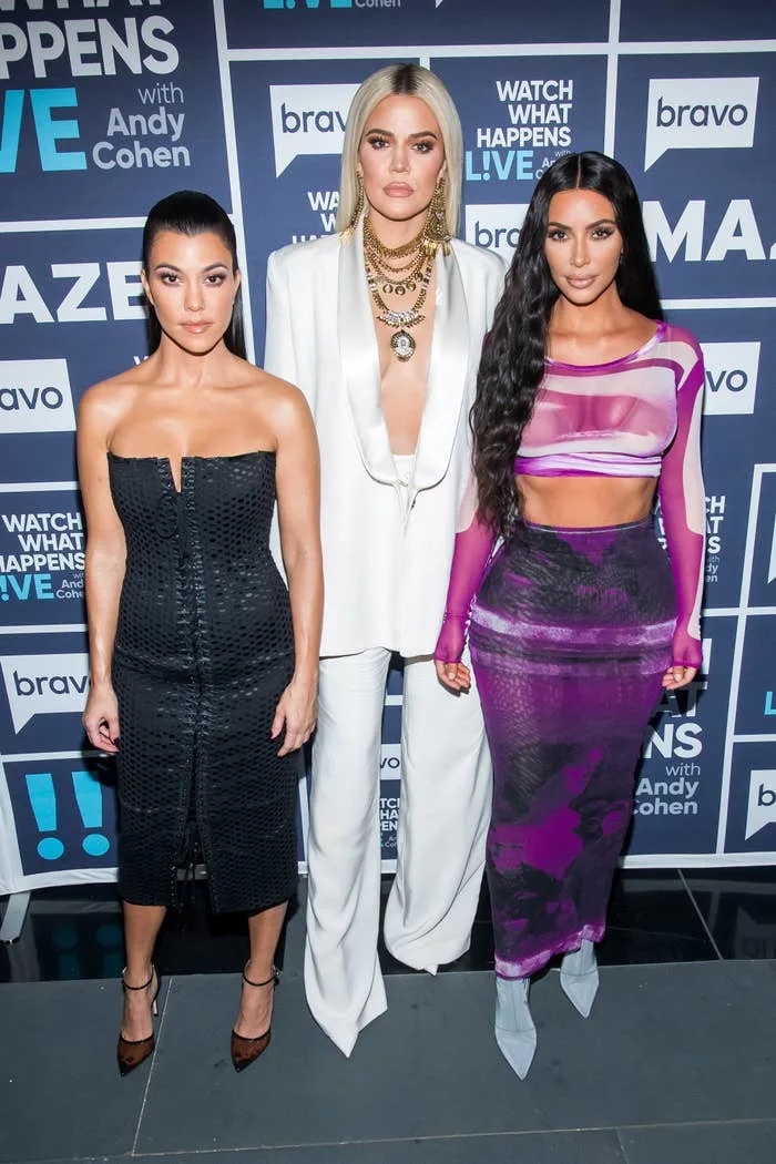 Kourtney, Khloe, and Kim Kardashian stand against a media event backdrop in stylish outfits