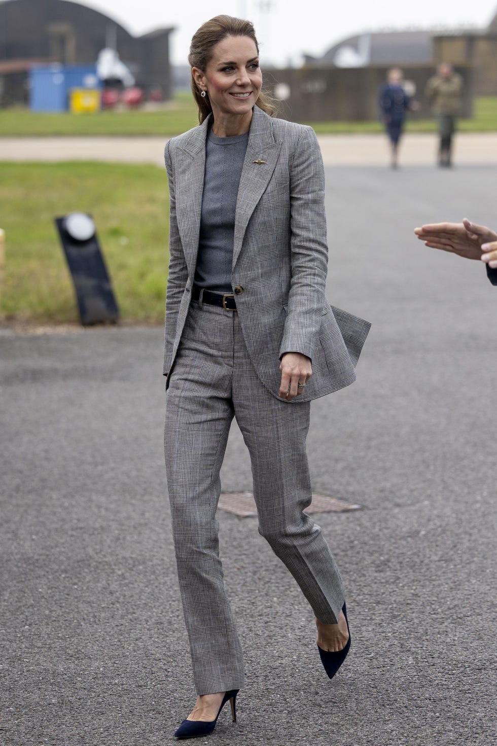 woman in tailored gray suit walking on tarmac while engaging with a man in a military uniform