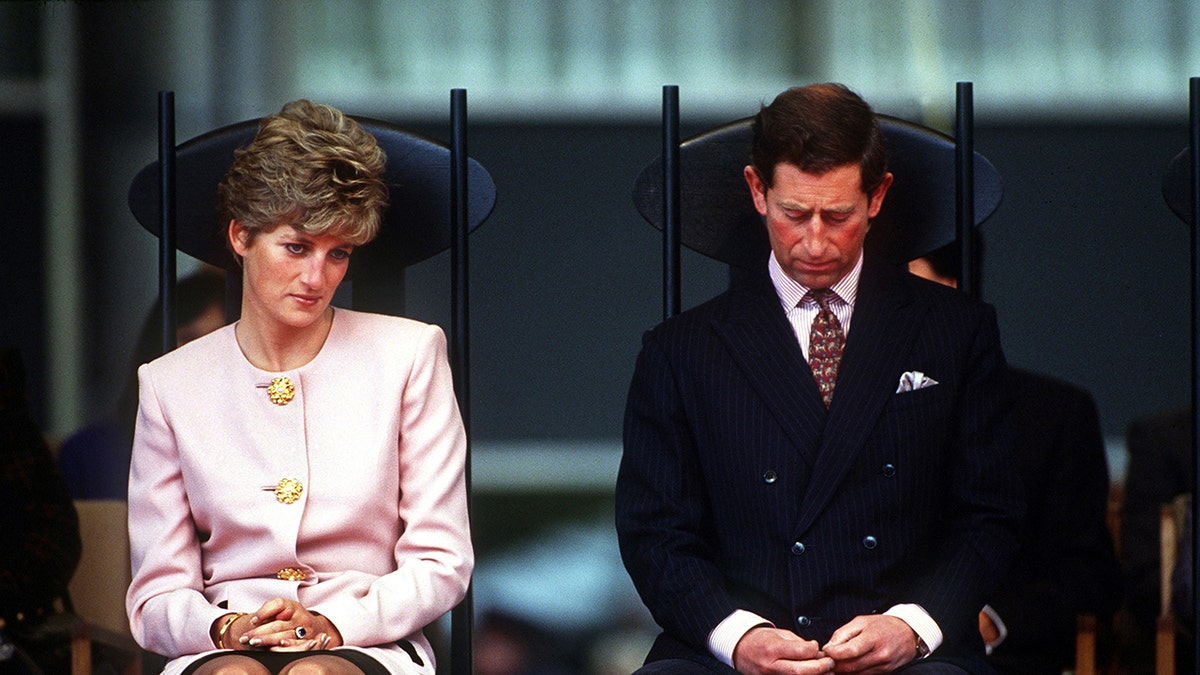 King Charles looking down in a black suit as Princess Diana in a pink suit looks on.