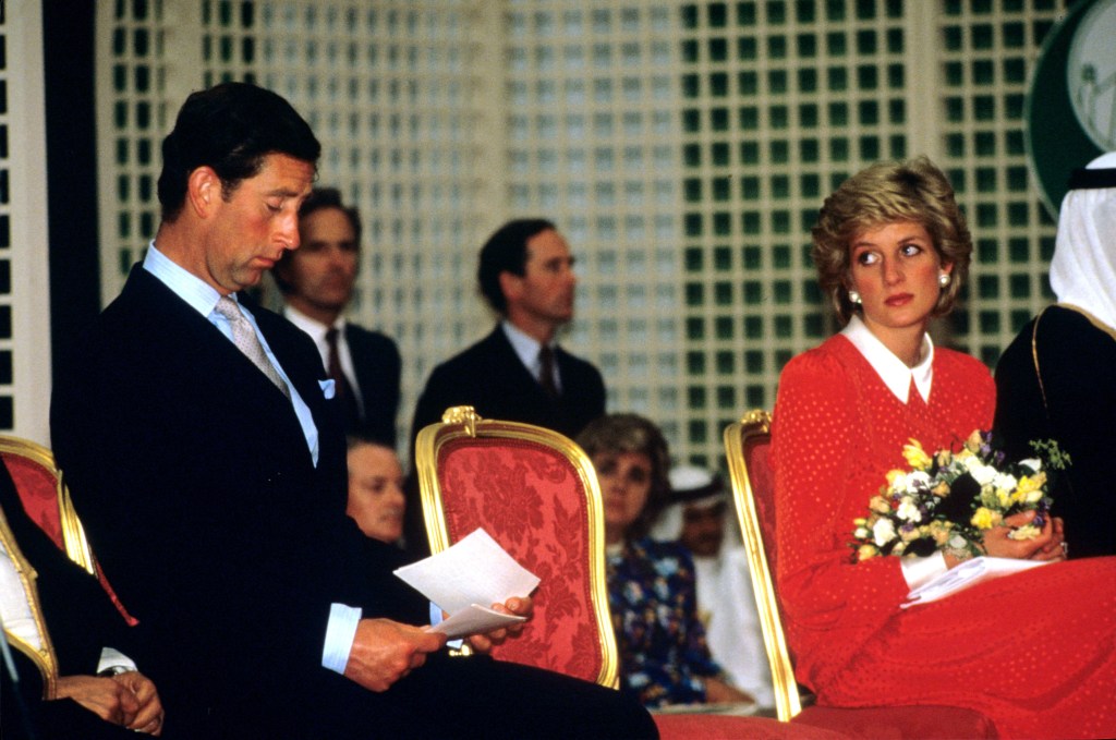 Prince Charles and Princess Diana seated away from each other. Princess Diana looks over forlornly at Prince Charles who is looking down.