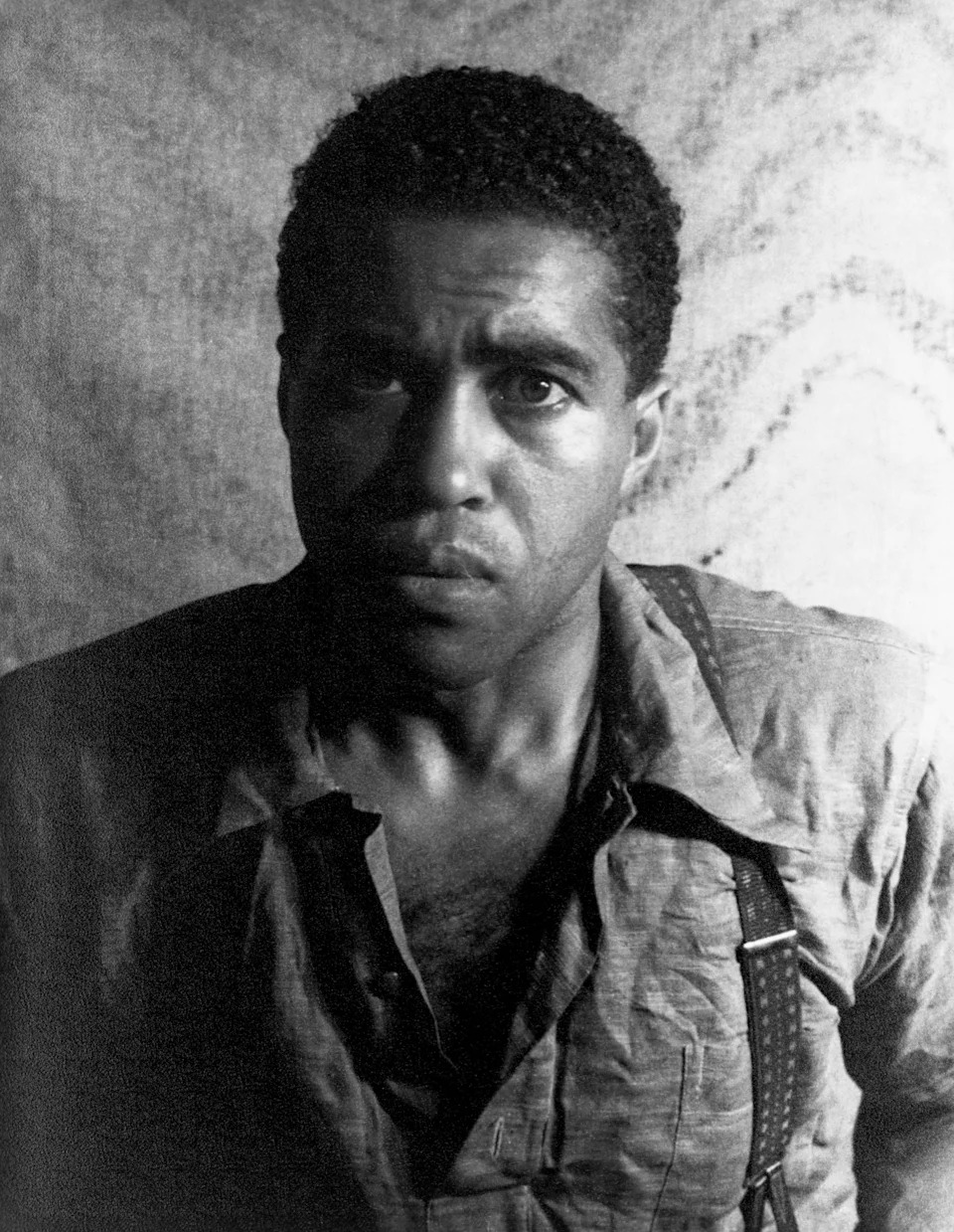 A person with a serious expression wearing a textured shirt and suspenders, looking directly at the camera against a plain backdrop