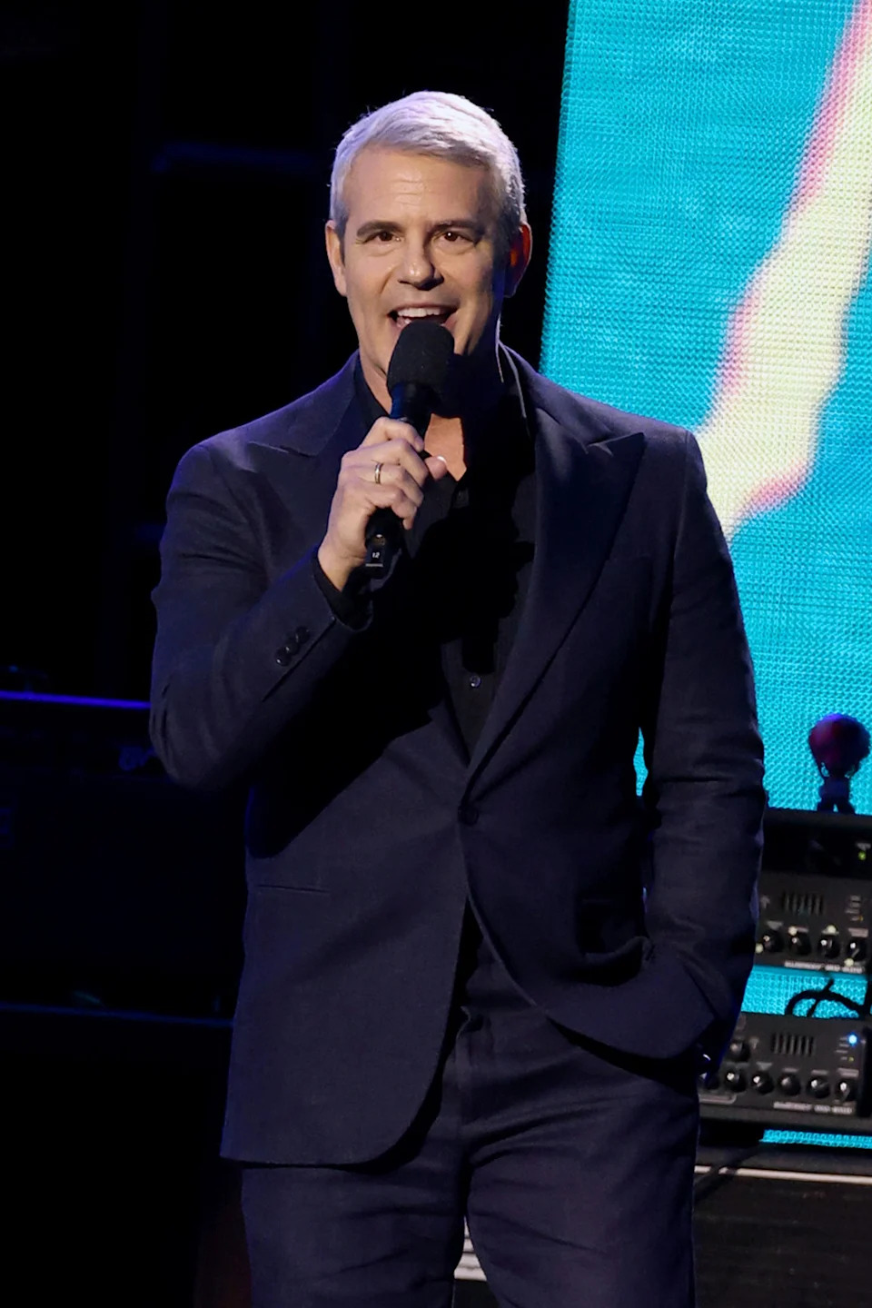 Andy Cohen in a formal suit holding a microphone on stage, speaking at an event