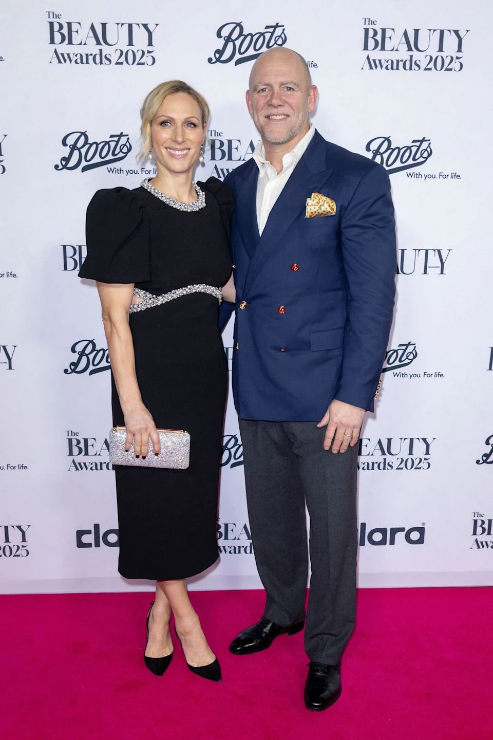 Zara Tindall in a black dress posing with Mike Tindall, wearing a blue blazer and gray pants, on a red carpet