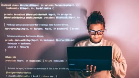 Getty Images A woman wearing glasses with her hair tied up looks smiling holding a laptop computer in her right hand whilst typing with her left. Programming code appears on the left hand side behind her as a back drop.