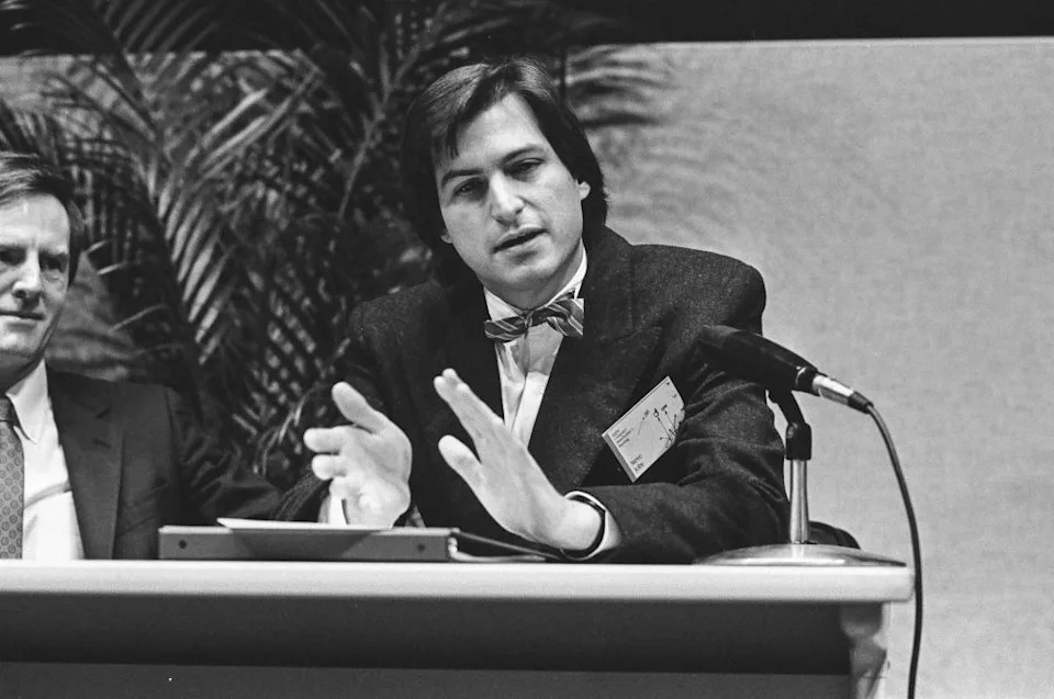 Steve Jobs, wearing a suit and bow tie, gestures while speaking at a panel discussion, with a microphone in front of him. Another man is seated beside him