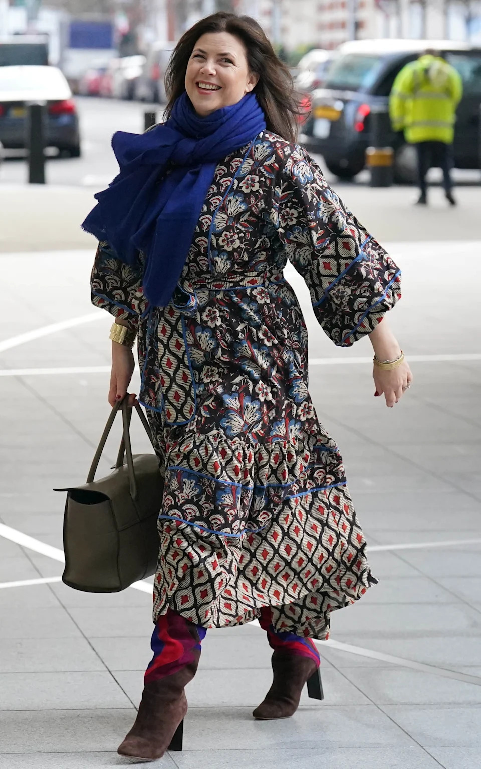 A smiling woman wearing a patterned dress, blue scarf, and brown boots walks on a street, holding a handbag