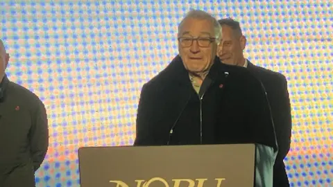 A man wearing a black jacket stands in front of a stand with a microphone. he is wearing glasses and has short grey hair. 