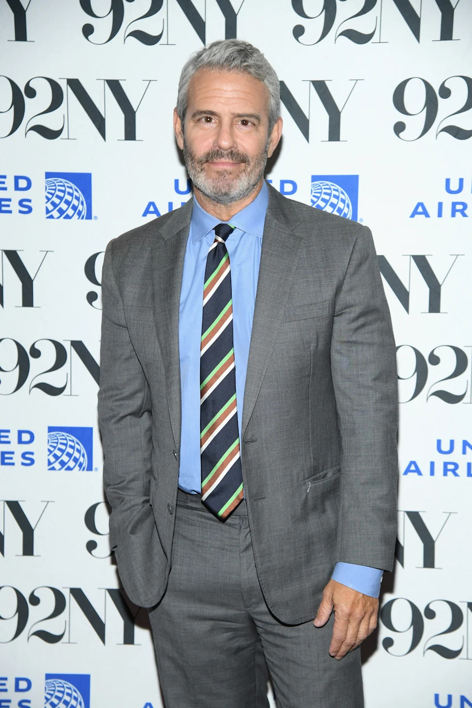 Andy Cohen in a suit with a striped tie at a 92NY event backdrop