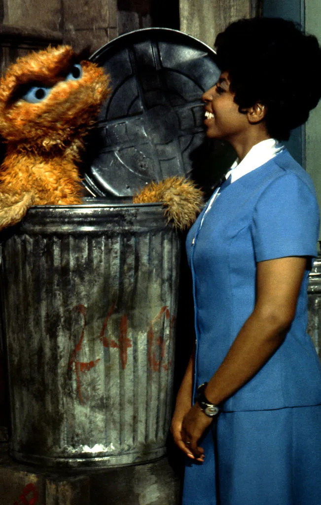 Oscar the Grouch in a garbage can talking to Loretta Long, who is wearing a blue dress with white accents