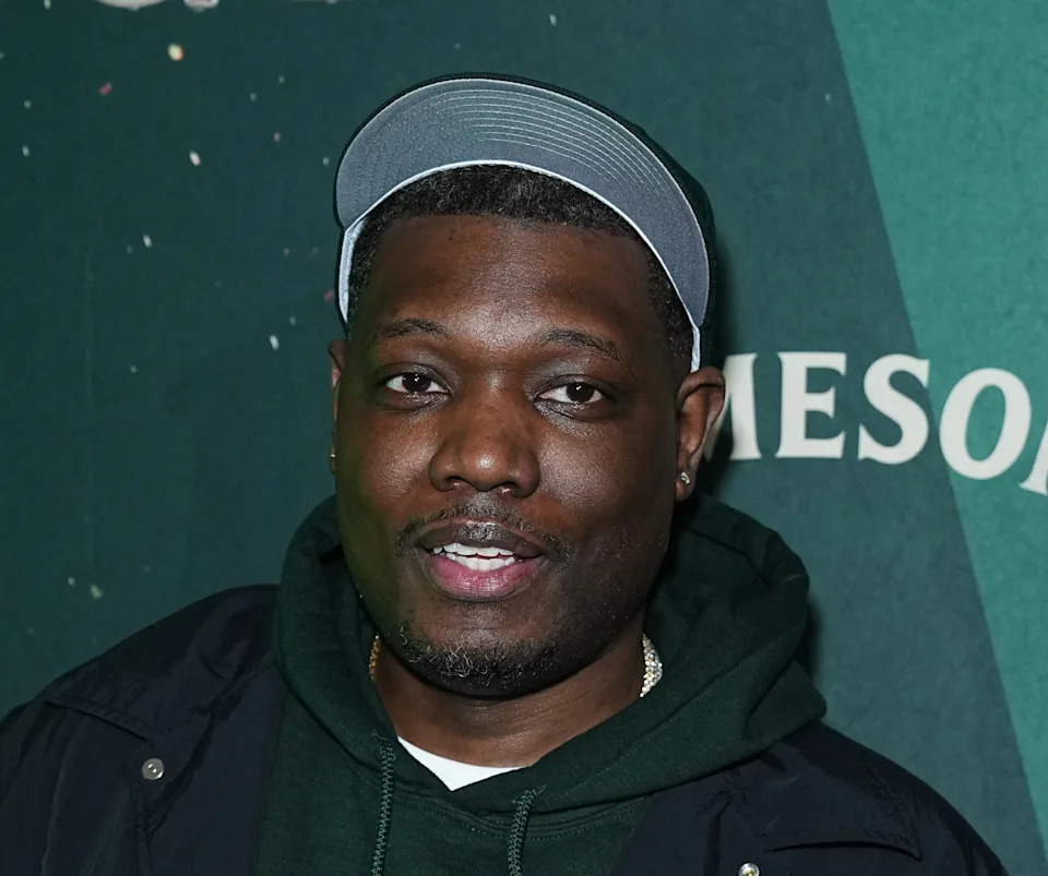 Person in casual attire with a jacket and hoodie, wearing a cap, posing at a public event