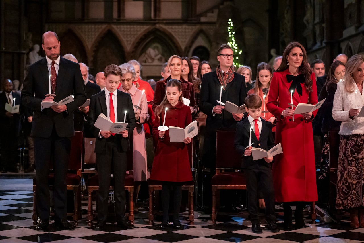 Adults and children stand in a row in front of chairs and holding candles in a large abbey.