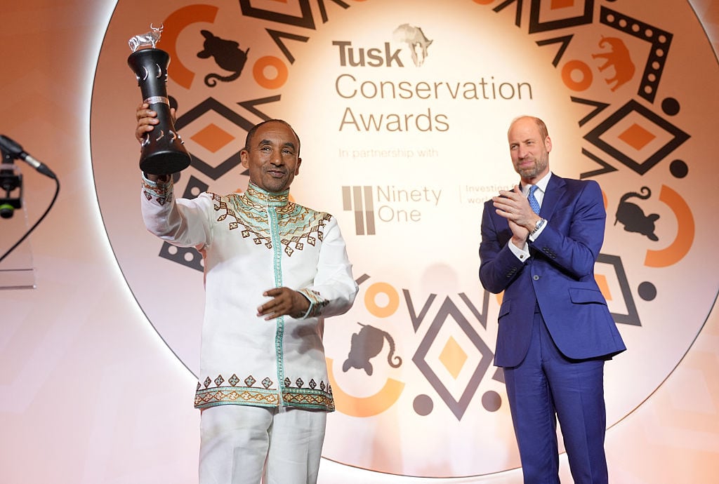 Two people stand on a stage as one claps and the other holds a trophy.