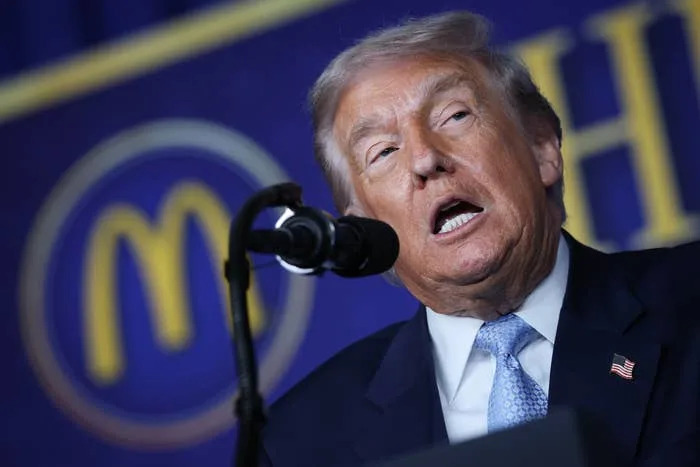 Donald Trump in a suit speaks at a podium with a microphone, appearing engaged