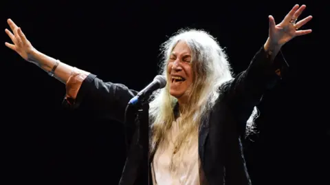 Getty Images Patti Smith with her arms outstretched and singing on stage with a black backdrop. Dressed in dark jacket with white shirt. 