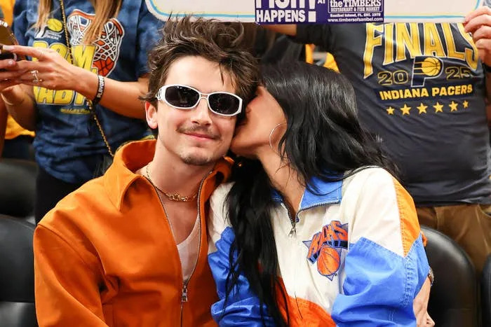 Timothée Chalamet and Kylie Jenner sit together; the man wears sunglasses and an orange jacket, while the woman wears a blue and white jacket, whispering to him
