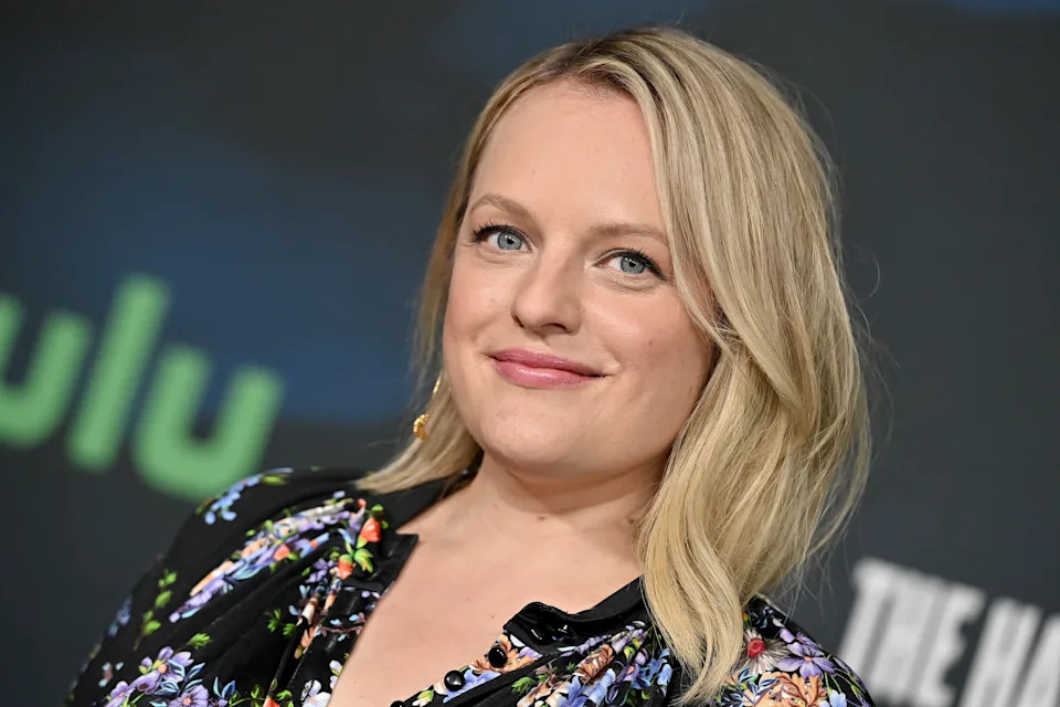 Smiling individual with shoulder-length blonde hair in a floral dress at a media event