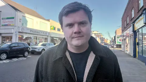 Councillor Aaron Skinner is outside on a street with cars on the road behind him. He has brown coat on and a black top