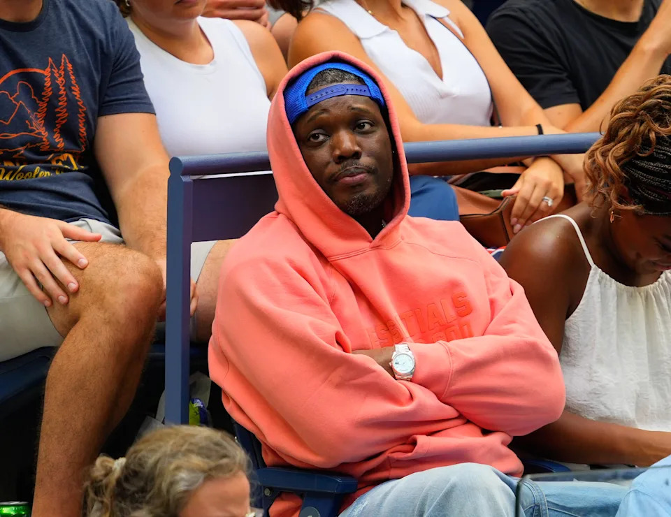 A person is sitting in a stadium, wearing a casual hoodie and cap, surrounded by other spectators. Their expression is thoughtful