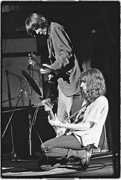 Jody Caravaglio Black and white photo shows Patti in the foreground bent down playing guitar - side profile looking down - wearing a white T-shirt and jeans.
Husband Fred is standing next to her playing guitar in a checked jacket.