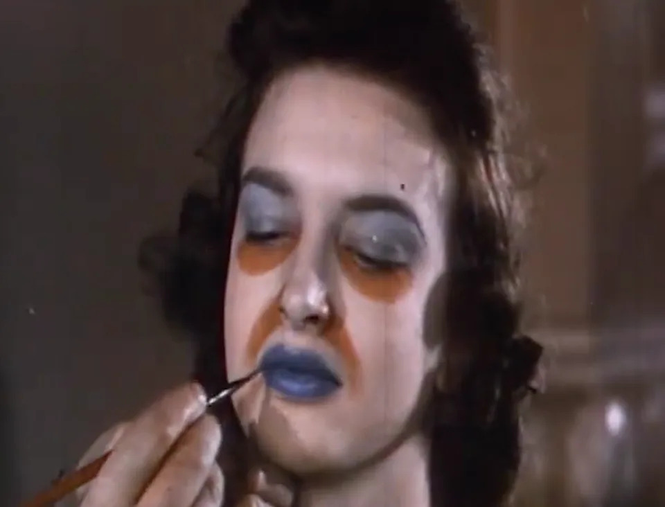 Person applying exaggerated makeup to a woman's face, giving a theatrical or artistic look, for a feature in a Rewind category article