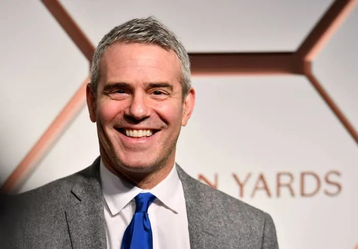 Andy Cohen smiles in a suit and tie at a formal event backdrop