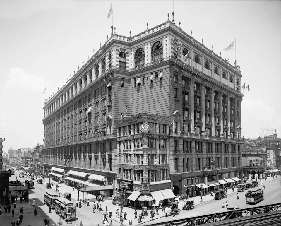Historic department store building with bustling street, vintage cars, and pedestrians. Ornate architecture and shop signs visible. Early 20th century vibe