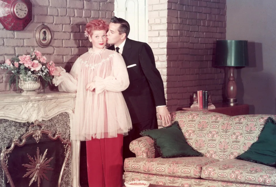 A person in a 1950s-style outfit is kissed on the cheek by another in a suit beside a vintage living room setting