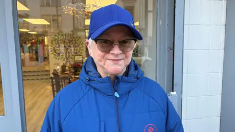 Robyn is wearing a navy cap and a navy coat. She has glasses on. She's standing on a street, with a business behind her.