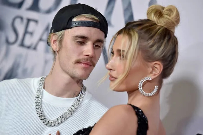 Justin Bieber in a cap, casual shirt, and chain and Hailey Bieber in a glam dress with large hoop earrings on the red carpet