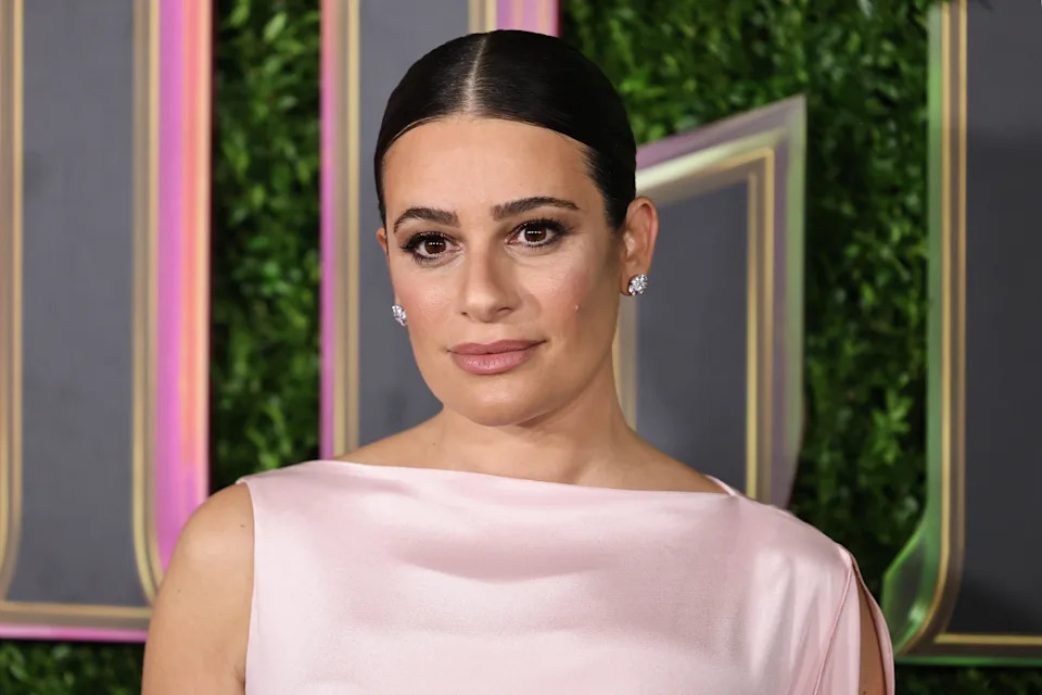 Person in elegant attire poses on a red carpet with a sophisticated, sleek hairstyle in front of a green backdrop