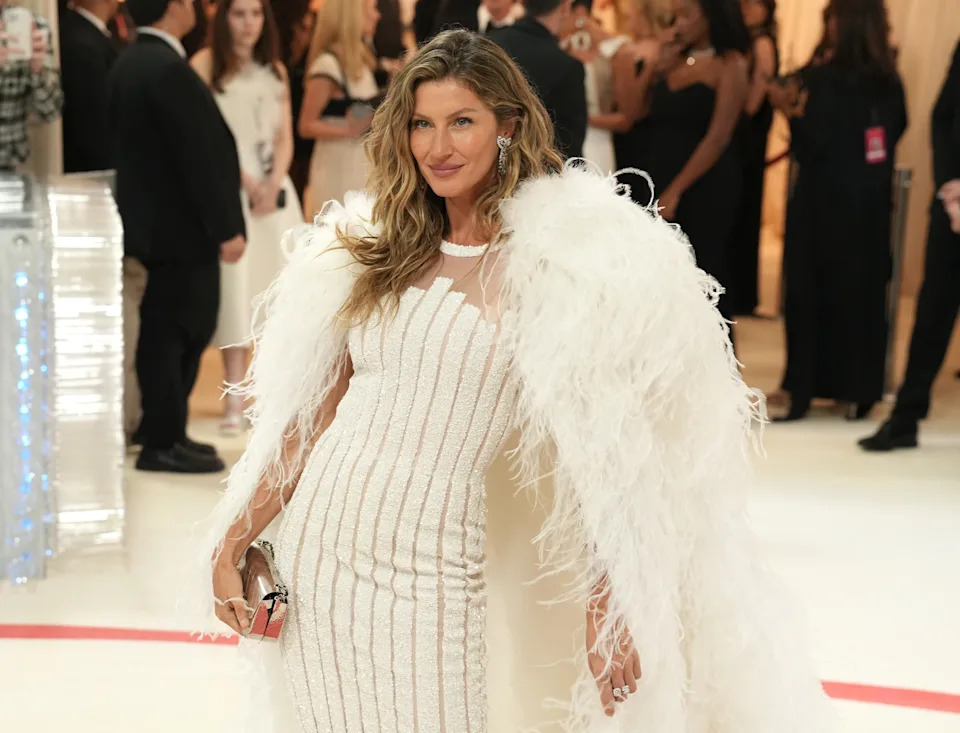 Person in an elegant white gown with a feathered cape, posing at a formal event with people in evening attire in the background