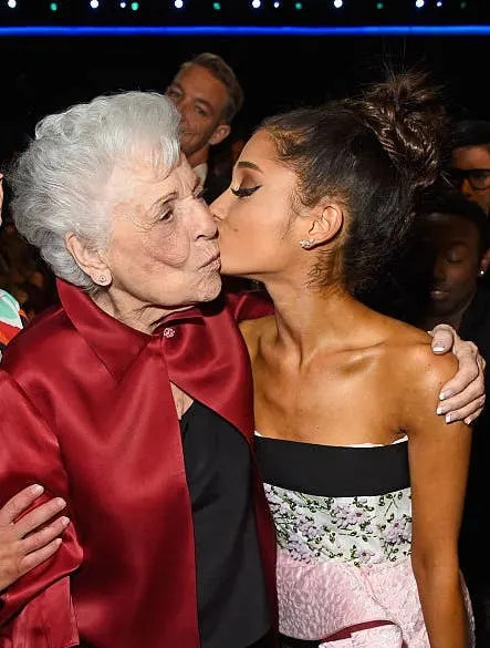 Three people posing at an event: one in a patterned blazer, an older person in a red shawl, and a woman in a strapless dress giving a kiss