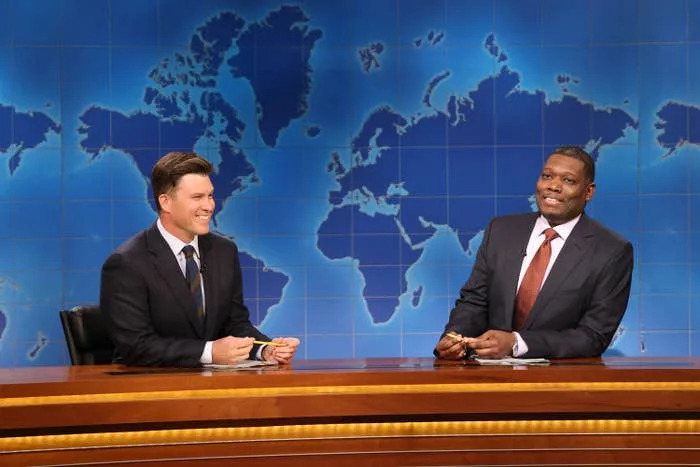 Two anchors in suits sit at a news desk on a set with a world map backdrop, likely during a televised segment