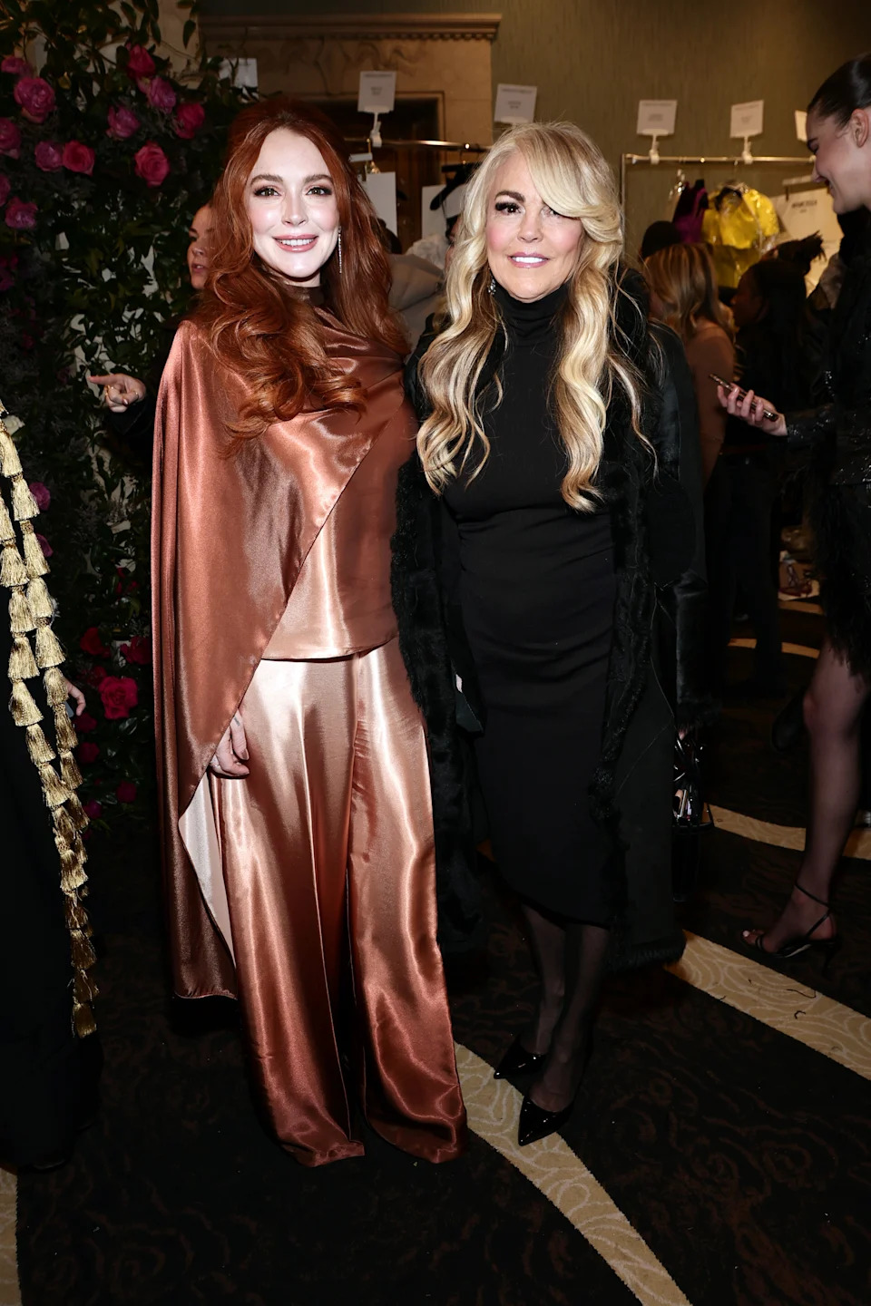 Two women pose together; one in a satin cape and pants, the other in a black dress with long sleeves. They are at a celebrity event