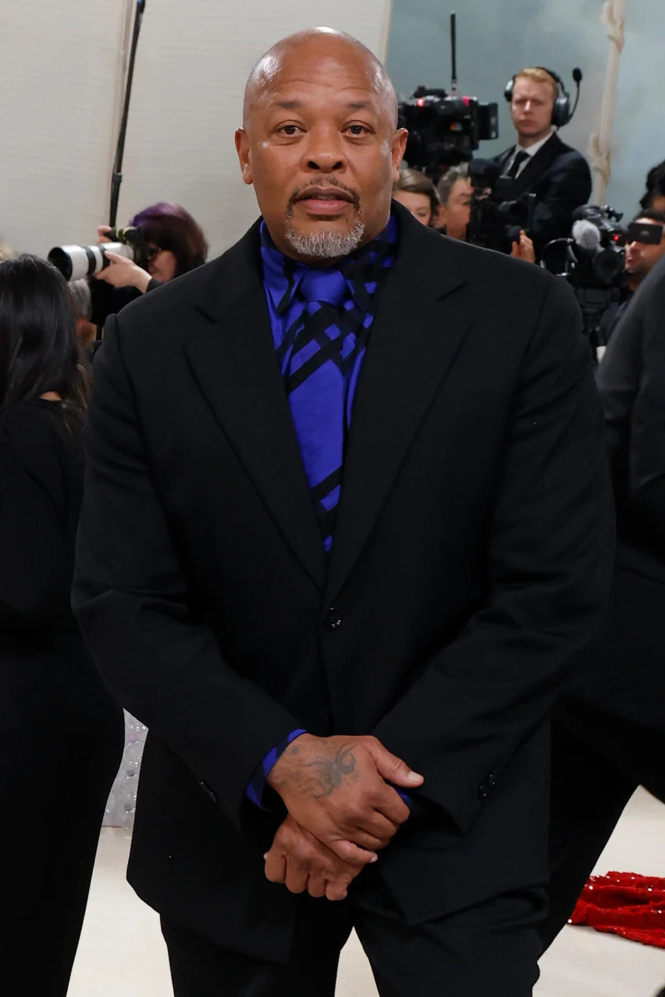 A person in a black suit with a patterned blue shirt stands on a red carpet at a celebrity event, surrounded by photographers