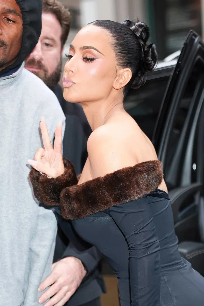 Kim Kardashian takes a photo with a fan giving a peace sign, wearing an off-shoulder dress with fur accents and sleek hairstyle