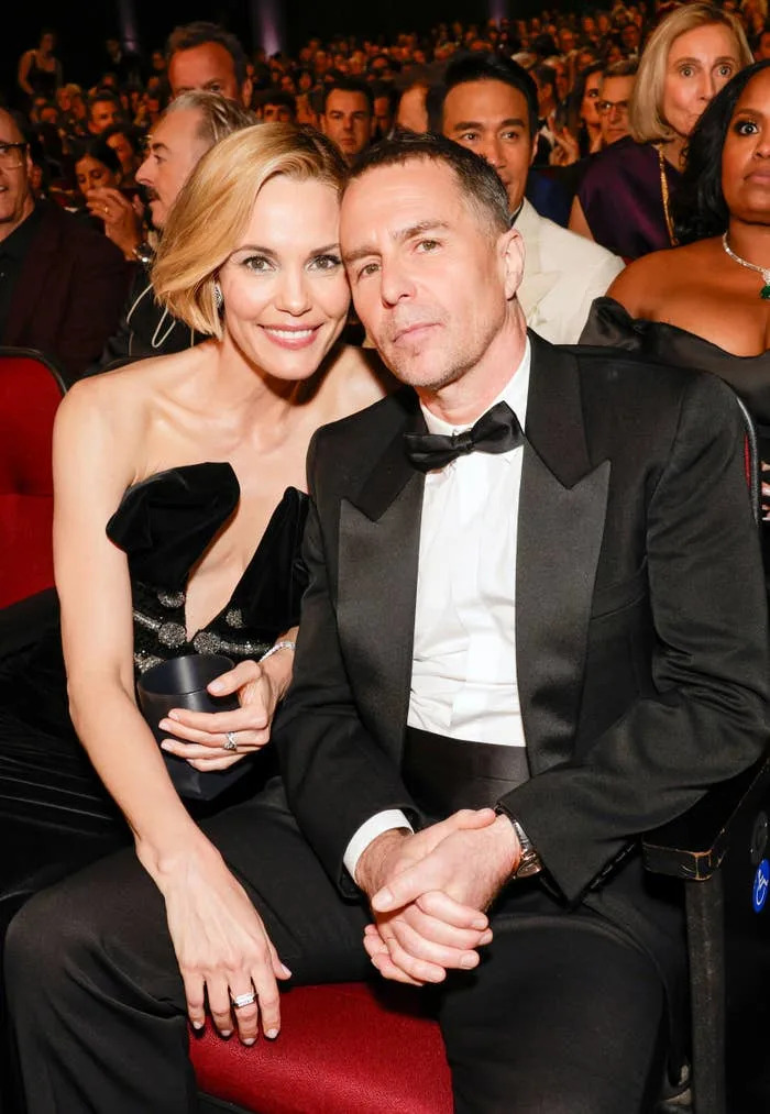Two individuals seated at an event, one in a black gown with a deep neckline, the other in a tuxedo with a bow tie, surrounded by an audience