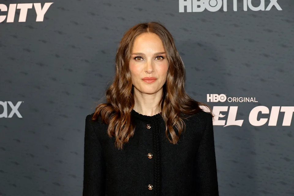 Celebrity posing at an event in a black, buttoned dress with wavy hair, standing against a backdrop with event branding