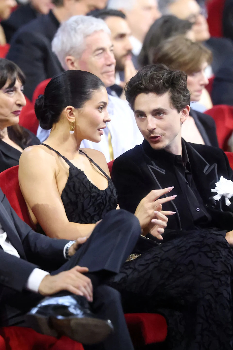 Kylie Jenner and Timothée Chalamet seated in elegant attire, engaged in conversation at a formal event. Others are seated behind them