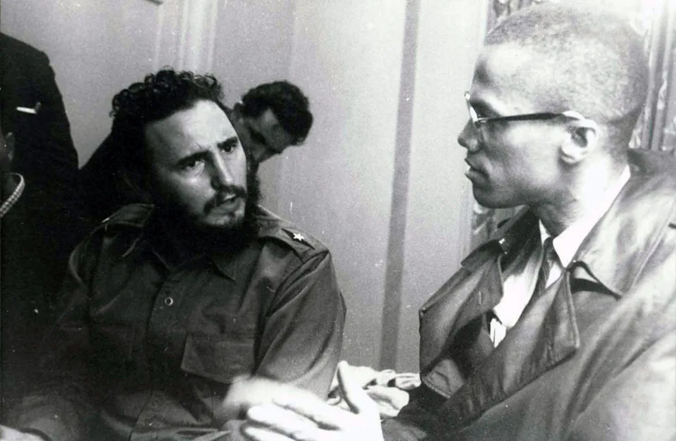 Two men in conversation, one wearing a military-style uniform, the other in glasses and a trench coat, engaged in a serious discussion