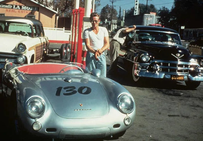 James Dean stands next to a Porsche 550 Spyder, with two classic cars parked nearby, at a gas station in the 1950s