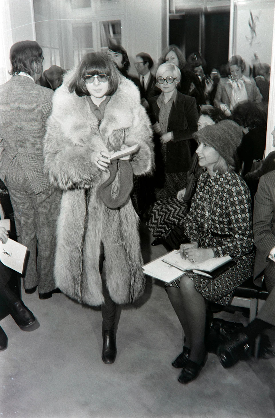 Person in a fur coat walking through a crowd, with people seated, writing on notepads in a retro indoor setting