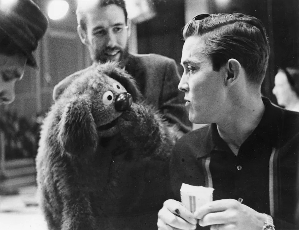 A man holds a puppet resembling a shaggy dog, engaged in conversation with another man holding a script, likely on a TV or film set