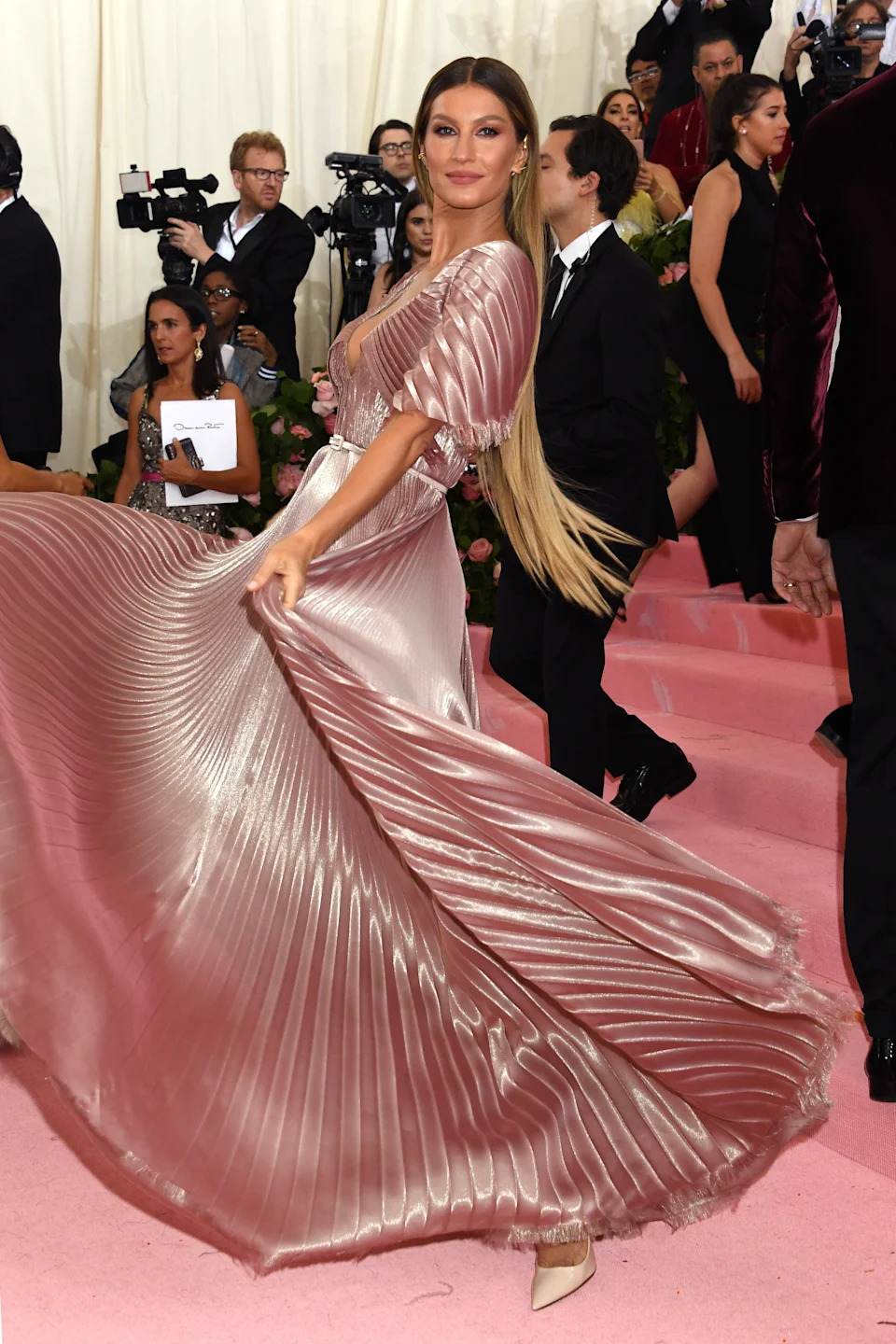 Celebrity poses in flowing pleated gown on red carpet event, photographers capturing the moment around them