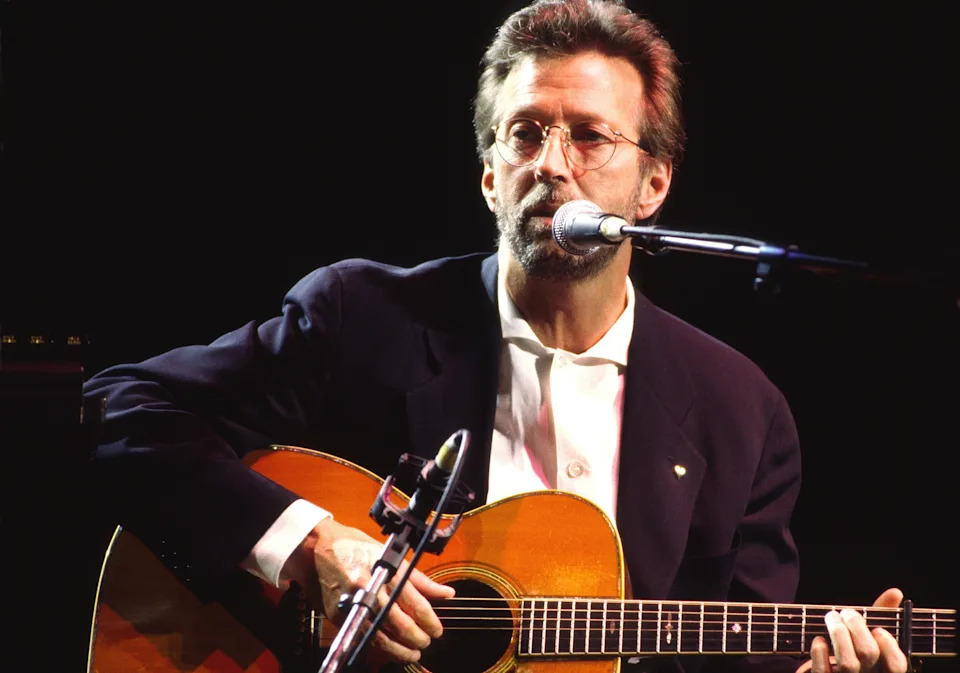 Musician on stage playing an acoustic guitar, wearing a suit and shirt, singing into a microphone